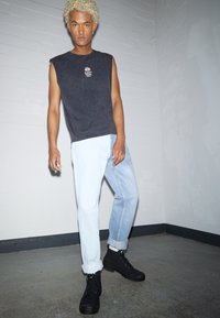 Černý bezrukávový top, světlé a tmavé modré džíny s ohrnutými nohavicemi, černé kotníkové boty, stojící na texturované podlaze s jednobarevným pozadím.