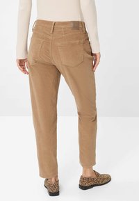Personne portant un pantalon en velours côtelé beige et des mocassins à imprimé léopard, se tenant contre un fond blanc uni, vu de dos.