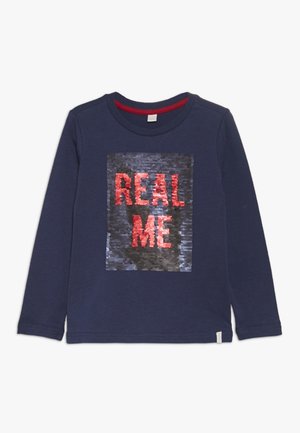Chemise bleu marine à manches longues avec un patch carré en sequins affichant les mots "REAL ME" en lettres rouges.