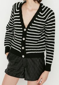 Gestreifter Cardigan in Schwarz-Weiß mit tiefem V-Ausschnitt, ausgestattet mit fünf dekorativen Knöpfen und gerippten Bündchen, kombiniert mit schwarzen wattierten Shorts.