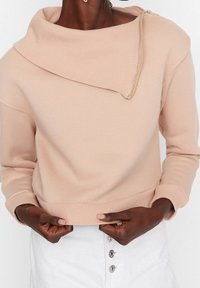 Sudadera de tela suave en tono rosa claro, con cuello en cowl y detalle de cremallera en el hombro. Puños y dobladillo ajustados, diseño sencillo.
