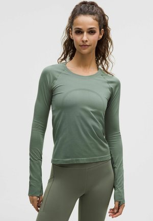 Kobieta o kręconych włosach, ubrana w dopasowaną zieloną sportową bluzkę z długim rękawem oraz dopasowane legginsy, stoi na jednolitym tle.