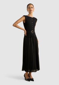 Musta kleit, millel on ilma varrukateta ülaserv ja nööpidega esikülg, kitsenev talje kullakettaga vööl, volditud seelik, koos lakkpindadega kontsakingadega.