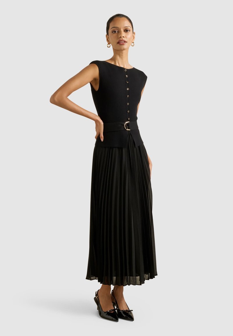 Musta kleit, millel on ilma varrukateta ülaserv ja nööpidega esikülg, kitsenev talje kullakettaga vööl, volditud seelik, koos lakkpindadega kontsakingadega.