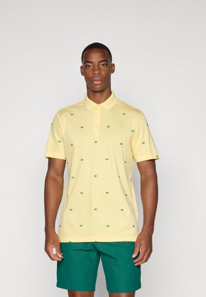 Yellow polo shirt with small green leaf patterns, short sleeves, and a collar. Worn with dark green shorts. Smooth fabric texture.