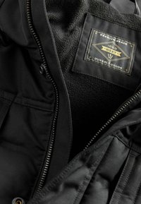 Schwarze Jacke mit einem weichen, strukturierten Innenfutter; verfügt über ein auffälliges Etikett, einen Reißverschluss und detaillierte Nähte entlang der Nähten.