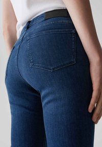 Jeans en denim bleu foncé avec une coupe ajustée, une taille mi-haute, des poches arrière au design classique et une étiquette en cuir noir à l'arrière.