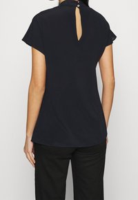 Haut noir à manches courtes avec un détail dos en trou de serrure et une fermeture à bouton. Tissu doux et léger avec une coupe décontractée et un ourlet arrondi.