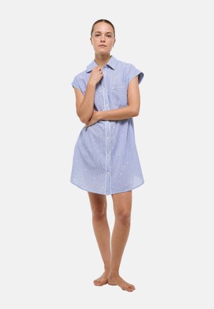 Femme debout pieds nus dans une robe à manches courtes, bleu clair, à boutons et à pois blancs, sur un fond uni.