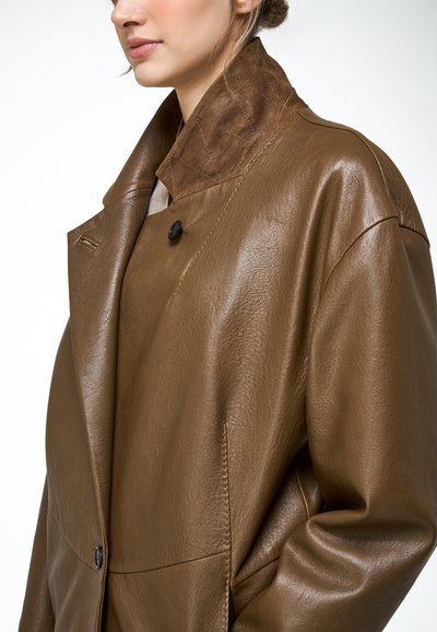 Kobieta ubrana w oversize'owy brązowy skórzany płaszcz z szerokim kołnierzem i czarnymi guzikami, pokazana od ramion do połowy tułowia.