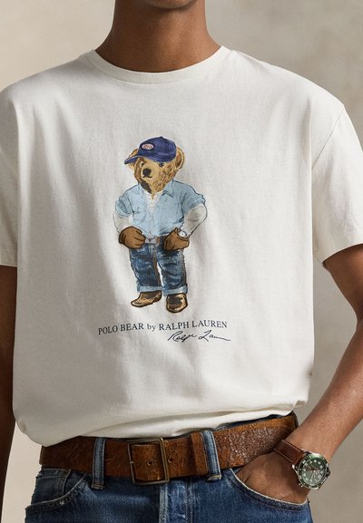 Fehér pamut póló, amelyen egy medve látható farmer ingben és kék farmernadrágban, kék sapkával és barna csizmával. Látható még egy barna öv és karóra is.