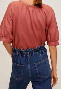 Blusa di lino color ruggine con maniche a sbuffo, abbinata a pantaloni in denim blu a vita alta con una fascia elastica arricciata in vita e tasche posteriori.
