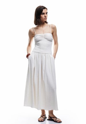 Vrouw draagt een mouwloze witte maxi-jurk met dunne bandjes en zwarte sandalen, staat met handen in de zakken, kijkt opzij.