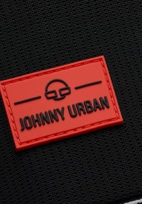 Piros gumi címke, amelyen a "JOHNNY URBAN" felirat domborítva szerepel egy texturált fekete háttéren. A szöveg felett szemüveg grafika található.