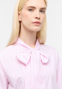Blusa rosa chiaro con righe bianche verticali, caratterizzata da un grande dettaglio a fiocco al collo e una texture liscia e raffinata.
