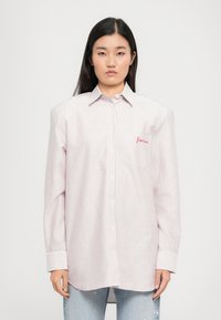 Mulher com cabelo longo e escuro, vestindo uma camisa branca oversized com finas riscas vermelhas e texto bordado no bolso do peito, combinada com jeans azul claro.