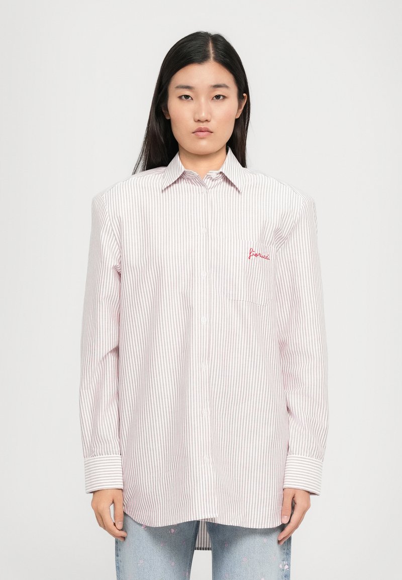 Mulher com cabelo longo e escuro, vestindo uma camisa branca oversized com finas riscas vermelhas e texto bordado no bolso do peito, combinada com jeans azul claro.