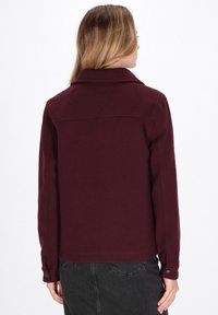 Burgundy takki, jossa on kaulus, napitettu hihansuu ja sileä rakenne. Sisältää takana ommellun sauman ja suoran leikkauksen.