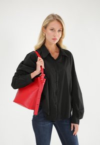 Sort langærmet skjorte med knapfront, parret med en tekstureret rød læderbucket taske med skulderrem. Modellen poserer med jeans.