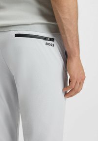Pantaloni sportivi grigio chiaro in materiale testurizzato, con una tasca con zip e un logo nero sul lato, accompagnati da un primo piano della mano e del tessuto.