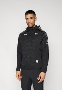 Veste coupe-vent noire avec un motif géométrique texturé, poches zippées et capuche. Comprend le logo UFC et des accents stylisés sur les manches.
