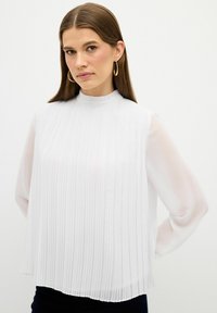 Weiße plissierte Bluse mit hochgeschlossenem Kragen und transparenten langen Ärmeln. Sie verfügt über ein fließendes Design und eine glatte Textur.
