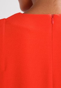 Červený oděv s hladkým, texturovaným materiálem. Má zadní zip s malým táhlem. Jednoduchý design a kulatý výstřih.