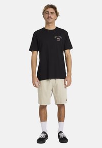 Zwarte T-shirt met korte mouwen met een kleurrijk logo op de linkerborst, gecombineerd met beige ribfluwelen shorts en zwarte sneakers.