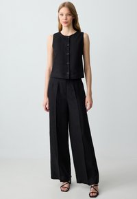 Gilet corto in lino nero con scollatura rotonda e chiusura frontale con bottoni, abbinato a pantaloni larghi neri e sandali neri con cinturini.