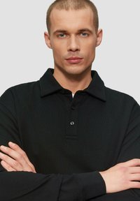 Schwarzes Poloshirt aus strukturiertem Stoff, mit Knopfleiste und Kragen. Das Modell steht mit verschränkten Armen vor einem neutralen Hintergrund.