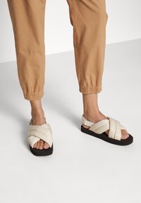 Beige padded sandals with crisscross straps and a black platform sole. The design features a slingback style with an adjustable strap.