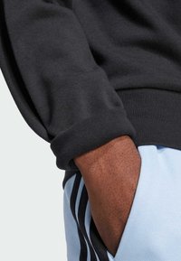 Sort sweatshirt med en struktureret overflade har manchetter. Lyseblå bukser med sorte striber er synlige, og der ses en lomme.
