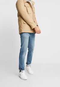 Beige, lätt jacka med knappstängning och framfickor, kombinerad med ljusblå jeans och vita sneakers.