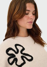 Light beige knitted sweater with a textured, oversized black flower design on the left side, featuring a round neckline and ribbed edges.