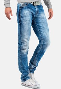 Jean en denim bleu avec un délavage usé, coupe slim et légères traces d'usure ; associés à des baskets montantes blanches.