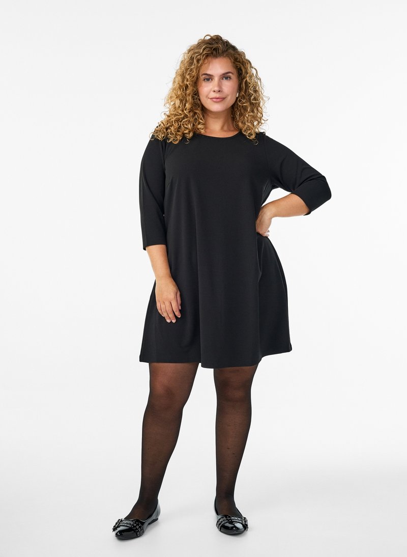 Donna con capelli ricci che indossa un vestito nero al ginocchio con maniche a tre quarti, collant neri e scarpe basse nere, in piedi con una mano sul fianco.