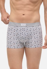 Hombre que lleva puesto un slip gris claro con pequeños patrones de pretzels azules y marrones y una cinturilla gris etiquetada como "Ysabel Mora".