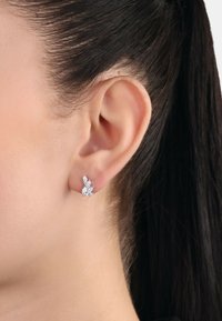 Små örhängen i silverfärg utformade som blad med flera klara ädelstenar, med en texturerad yta och en säker bakdel med stift.