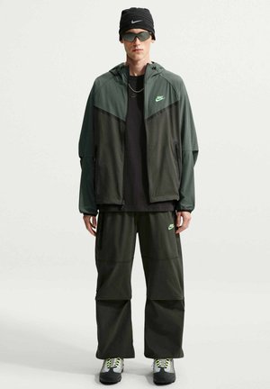 Mand iført mørkegrøn og sort Nike jakke og bukser, sort hue og solbriller, står mod en ensfarvet lys baggrund.