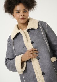 Chaqueta de ante sintético gris con un cuello y puños de borreguillo color crema, con botones negros y costuras blancas a lo largo de las costuras.
