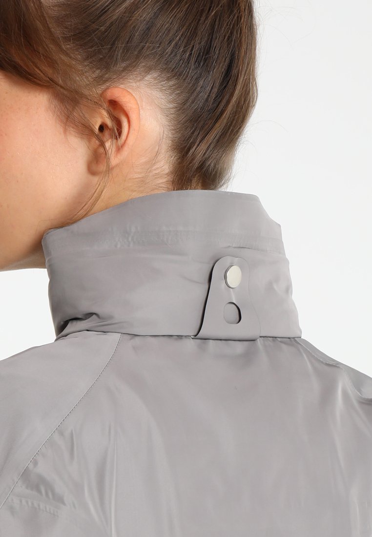 Graue wasserdichte Jacke mit hohem Kragen, ausgestattet mit einem Druckknopfverschluss und einer glatten Textur. Seitliche Nahtdetails sind sichtbar.