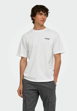 Giovane uomo con capelli ricci che indossa una t-shirt bianca a maniche corte con il testo "modul" e pantaloni cargo grigio scuro, guarda di lato.