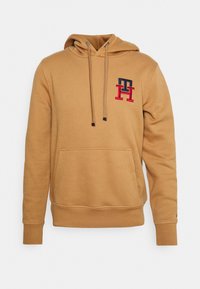 Sudadera color beige con un bolsillo frontal, capucha con cordones y un logo bordado en rojo, azul y blanco en el pecho. Hecha de suave tela de algodón.