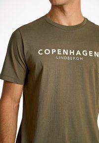 Camiseta de algodón verde oliva con mangas cortas, que presenta una etiqueta impresa en blanco que dice "COPENHAGUE" y "LINDBERGH" en el frontal.