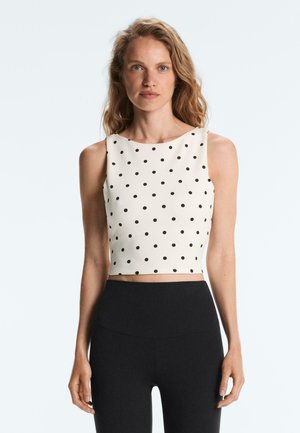 Donna che indossa un top corto bianco senza maniche con pois neri e pantaloni neri a vita alta, in piedi davanti a uno sfondo chiaro.