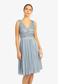 Robe bleu clair avec une jupe plissée, des détails en dentelle sur le corsage et un décolleté en V. Dotée de bretelles larges et d'une texture lisse.