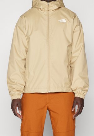 Veste à capuche beige de The North Face avec fermeture éclair frontale portée par une personne en pantalon orange, montrant des poignets élastiques et le logo sur la poitrine.