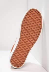 Semelle en caoutchouc orange texturée avec une adhérence à motif losange, présentant un bord latéral blanc. La chaussure est positionnée sur un fond clair.