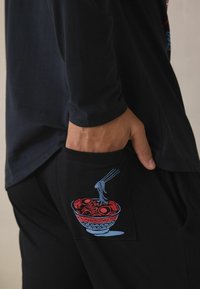 Zwarte longsleeve met een ontspannen pasvorm, gecombineerd met zwarte broek met een kleurrijke afbeelding van een kom ramen op de zak.