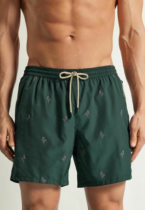 Mann trägt dunkelgrüne Badeshorts mit kleinen pinken Flamingo-Mustern und beigem Kordelzug, steht mit entspannten Händen an den Seiten.